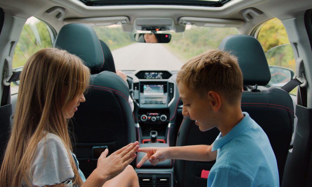 Back view of attractive friendly happy teen boy and girl which sitting at car beackseat and playing games during traveling together with their mom